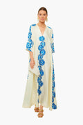 A woman wears a La DoubleJ silk cream maxi dress with hand-printed daisy motifs, paired with a white clutch, tan sandals, and gold earrings. Made in Italy.