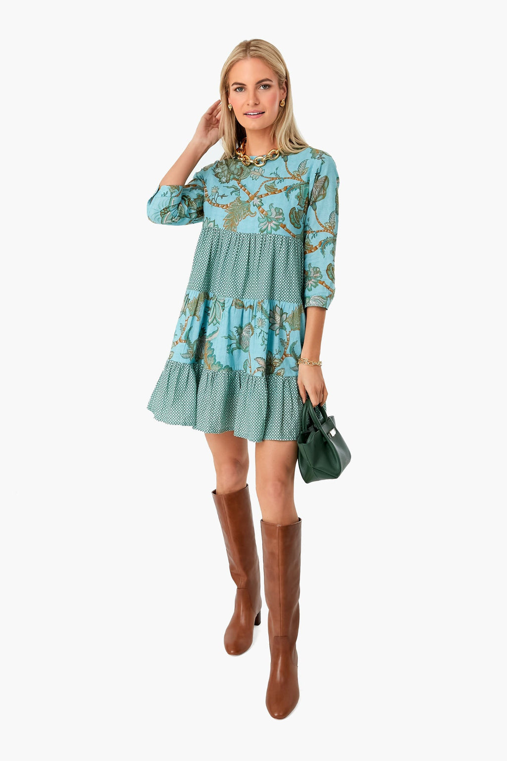 A woman wears the Ro¡¯s Garden Turquoise Camilla Rene Dress¡ªa blue patterned, tiered block-printed dress¡ªwith brown knee-high boots and a small green handbag.