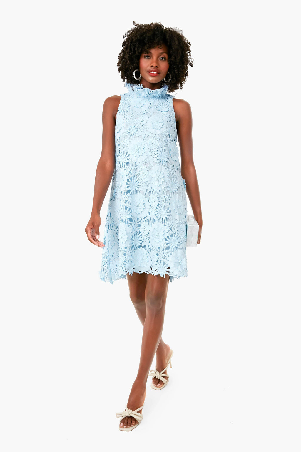 A woman in a Tuckernuck sleeveless light blue lace swing dress with a ruffle stand collar and open-toe bow heels walks forward, carrying a small clutch.